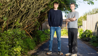 Two young men stand on a paved path between dense green bushes and a wooden fence, with one wearing a black sweater and jeans and the other in a gray shirt and black pants.