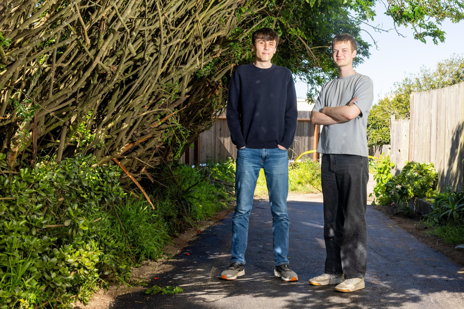 Two young men stand on a paved path between dense green bushes and a wooden fence, with one wearing a black sweater and jeans and the other in a gray shirt and black pants.