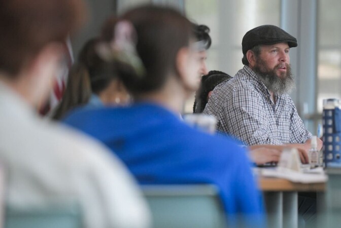 A man wearing a cap and plaid shirt is pictured in profile. He is seated, the backs of several people are pictured in the foreground