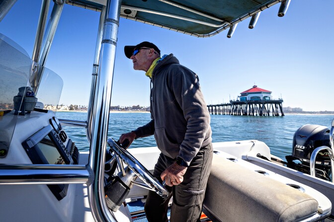 A man wearing black pilots a boat. Behind him, a pier juts into the ocean from a distant beach.