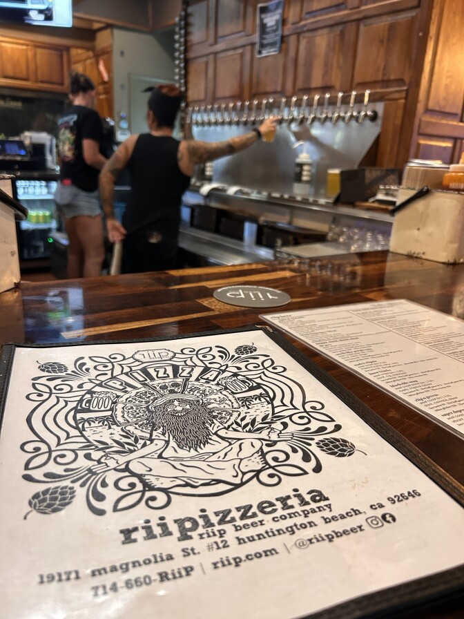 A woman pulls a tap behind the bar; the focus is on menu that says "Riipizzeria" on the bar.