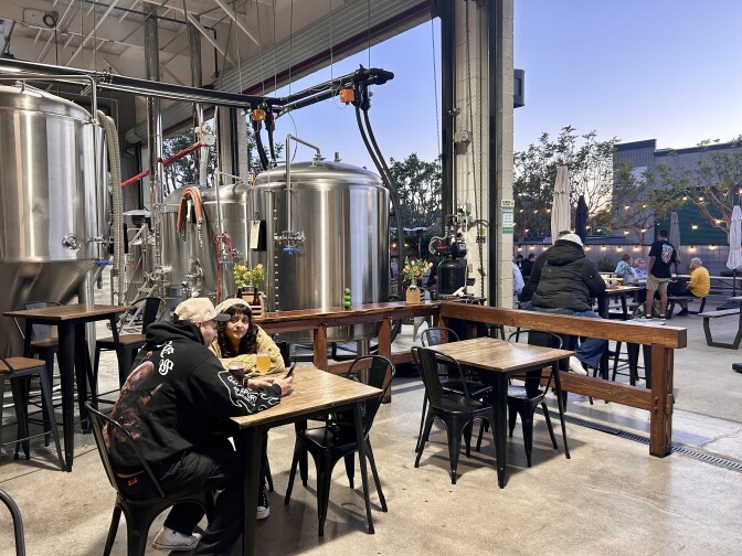 A couple sits at a table drinking beer in a room open to a patio with more people at tables, and large brewing vats in the background.