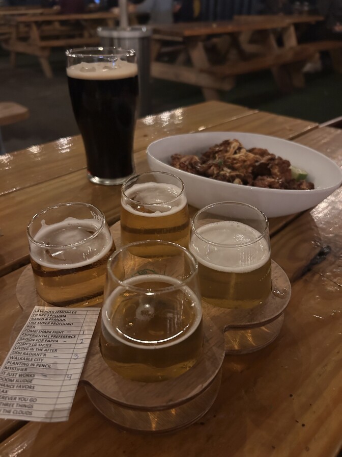 A flight of four beers on a table along with a bowl of food and another dark beer.