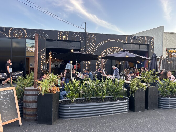 An outdoor patio with plants, black umbrellas and people sitting at tables, with a black building with gold patterns in the back.