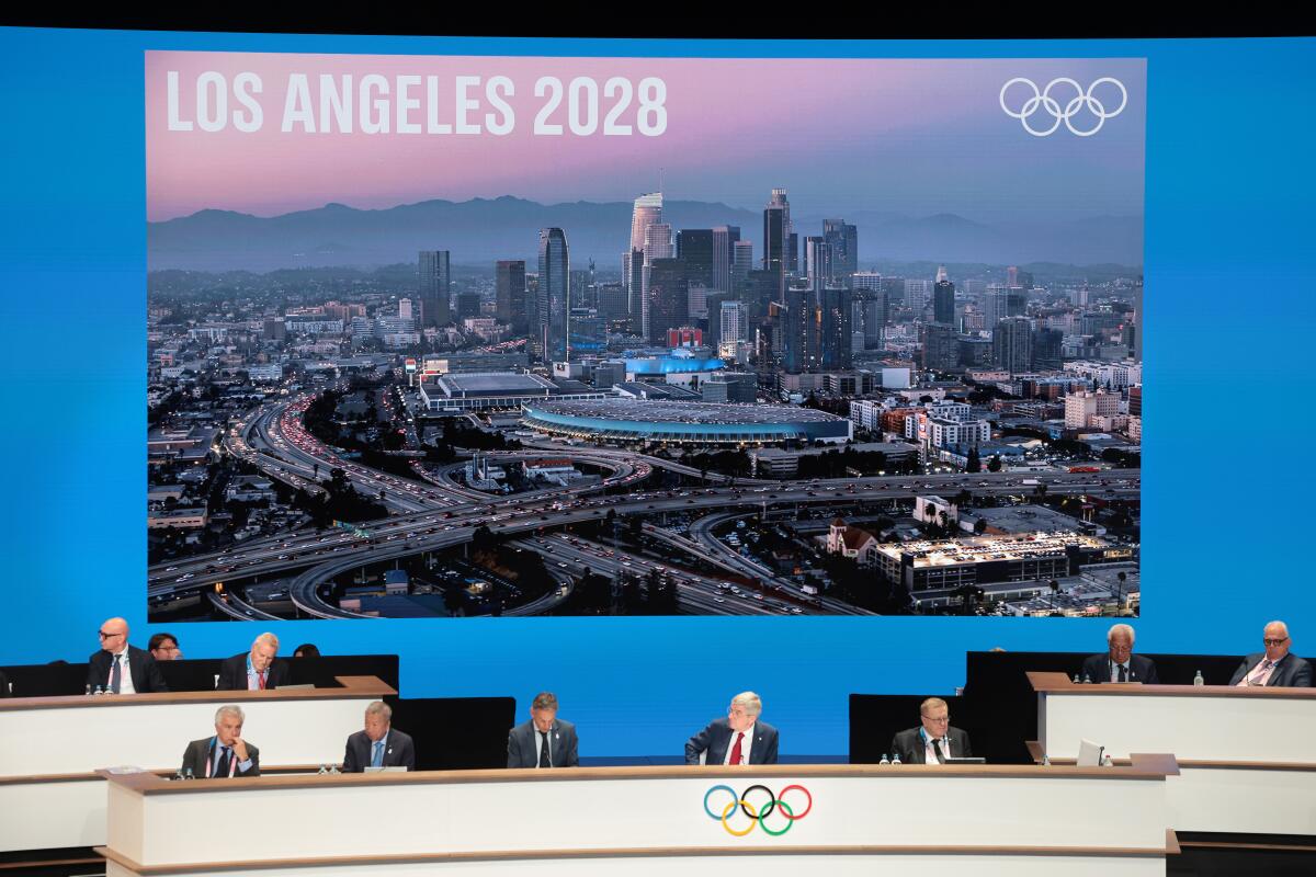 Dignitaries sit at the dais with the Olympics ring logo; behind them, a projection of downtown Los Angeles