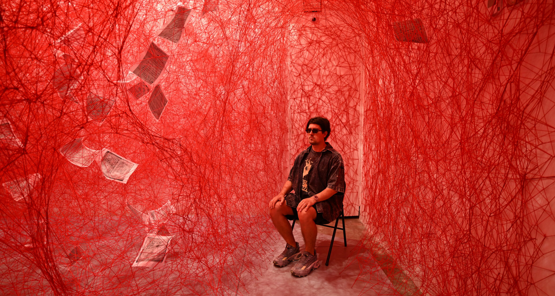 A person sits on a chair in a room filled with dense red string webs and suspended sheets of paper, under red lighting.