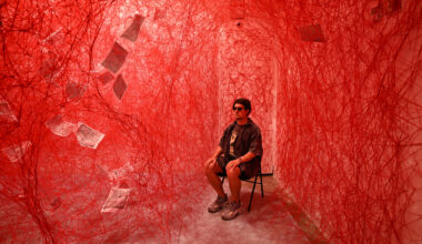 A person sits on a chair in a room filled with dense red string webs and suspended sheets of paper, under red lighting.