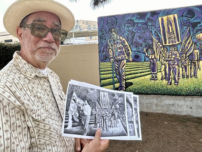 A male presenting person wears glasses and a hat. He holds sheets of paper.
