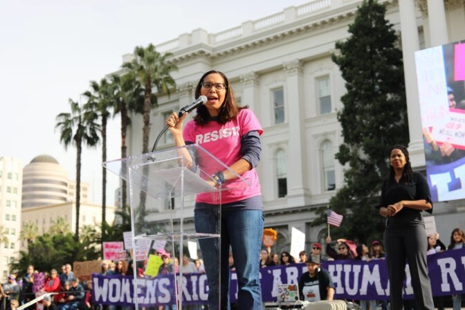 A woman in a pink shirt stands in front of a podium. She is speaking into a mic. She is wearing a pink shirt that has the text "RESIST" printed on it in white. Behind her, a crowd of people stand holding a banner that says "Women's Rights are Human Rights."