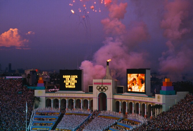 A stadium is full with audience members and two large scoreboard screen. The message on one of them reads: 'Good luck to the athletes of the world'. Purple smoke rises in the distance.