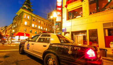 A black and white police car with flashing lights is parked on a street corner in a brightly lit city area with pagoda-style architecture at dusk.