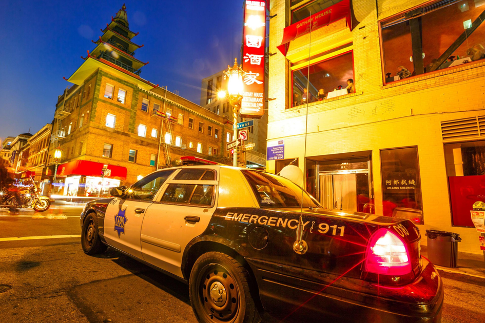A black and white police car with flashing lights is parked on a street corner in a brightly lit city area with pagoda-style architecture at dusk.