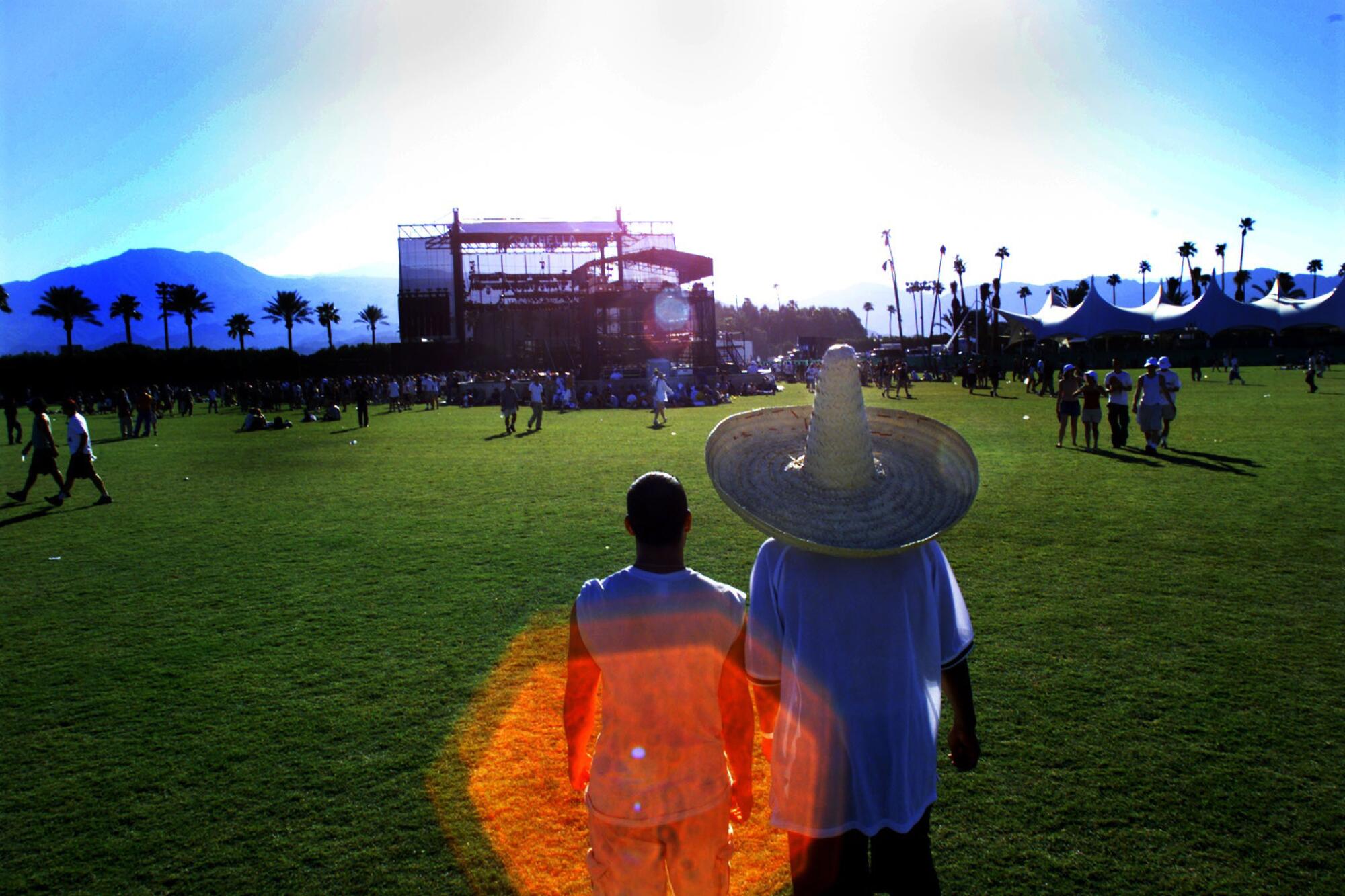 Two men walk on a sunny field with a stage and palm trees ahead of them. One is wearing a sombrero.