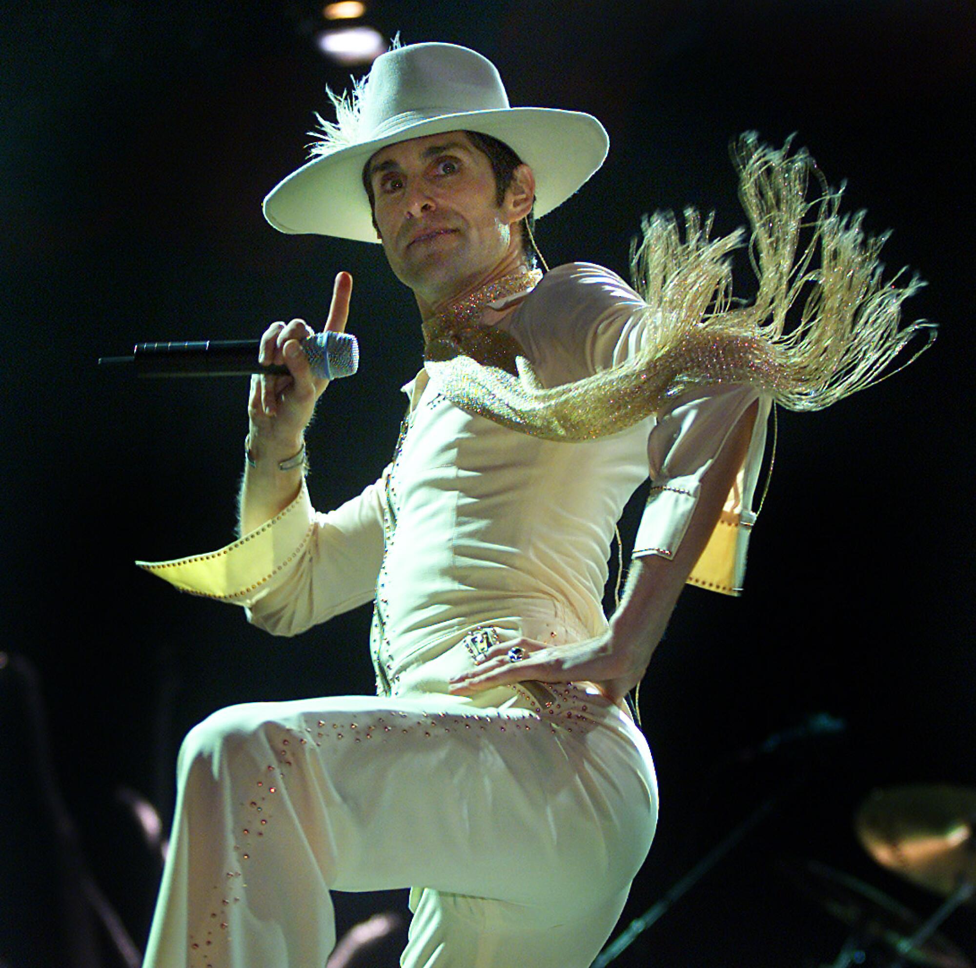 Perry Farrell holds a microphone while wearing a white flowy outfit with fringe and a large hat