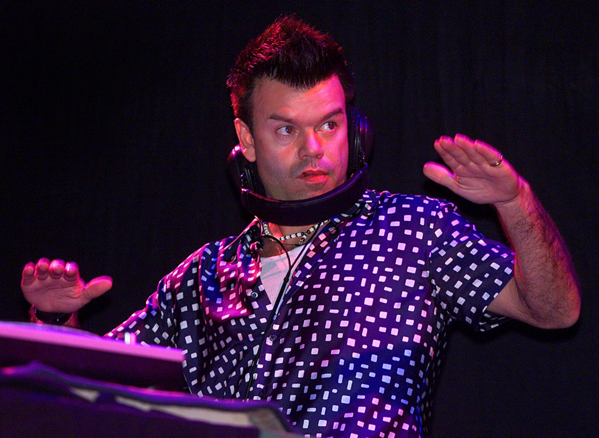 DJ Paul Oakenfold wears headphones around his chin and has his palms raised and facing down