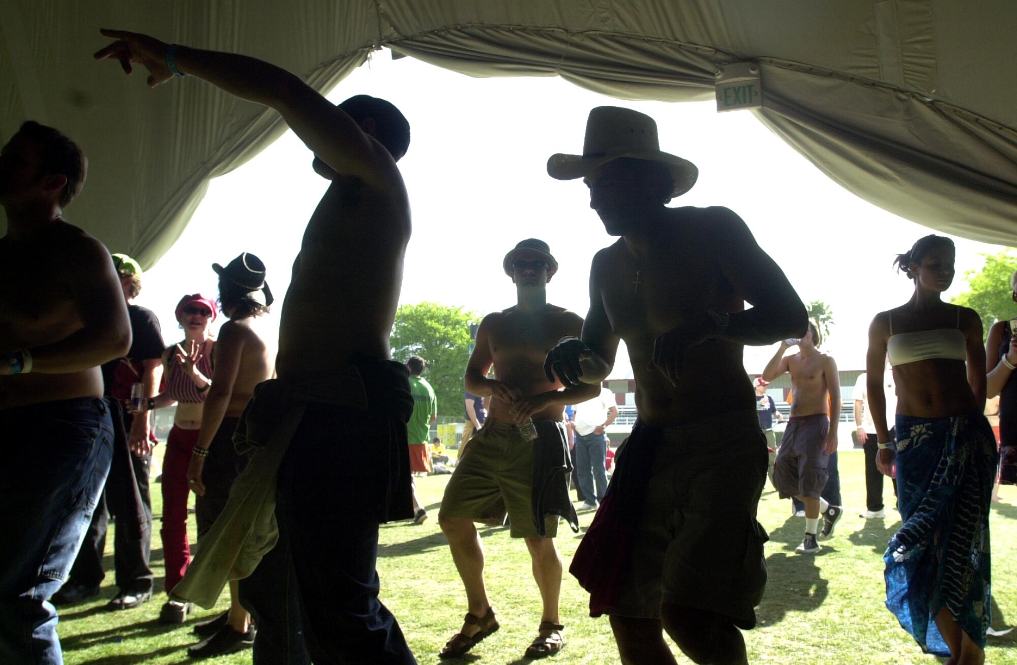 People dance in a tent in the bright sunlight on a field.