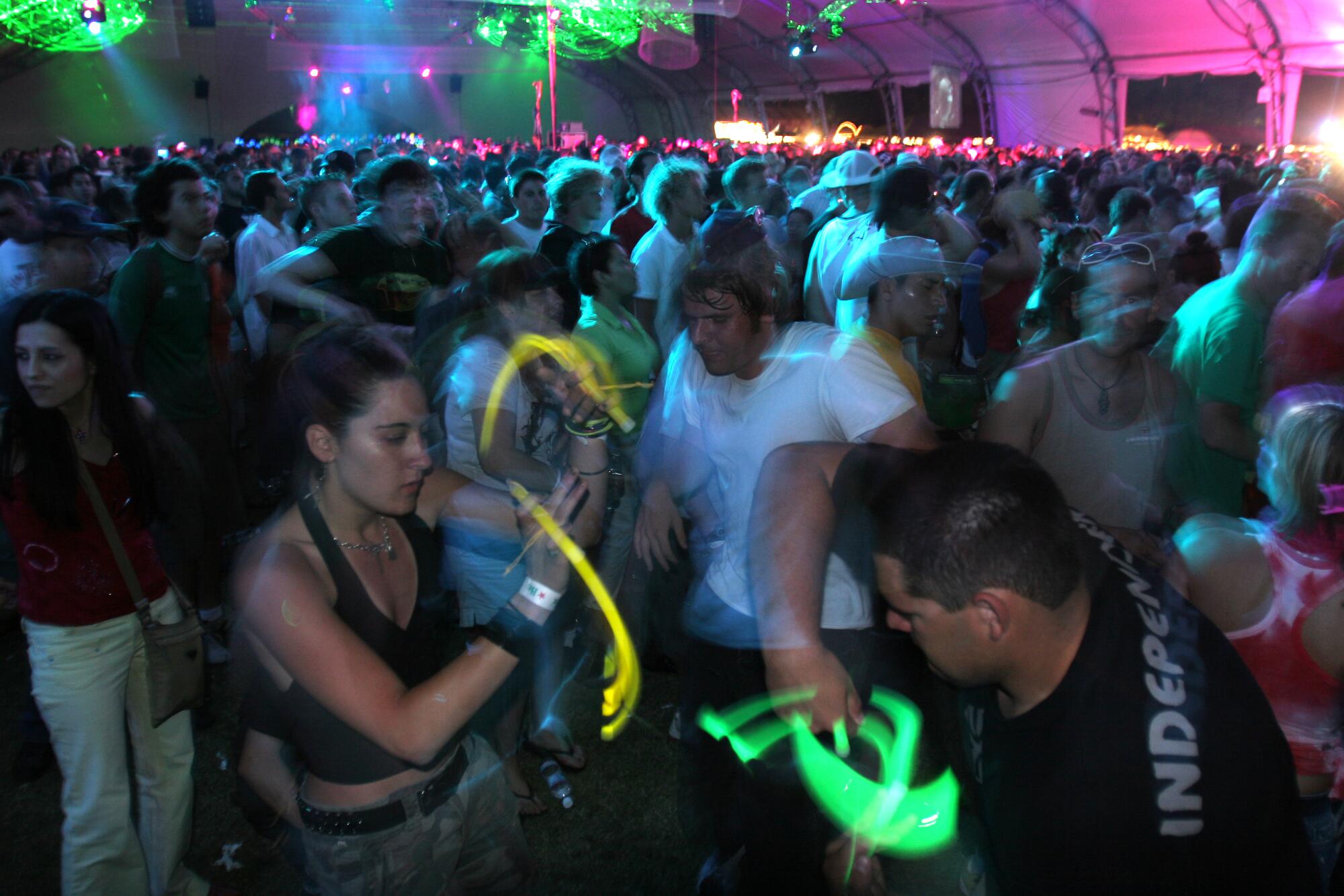 Thousands of people dance, including some with glow sticks, under a large tent at night