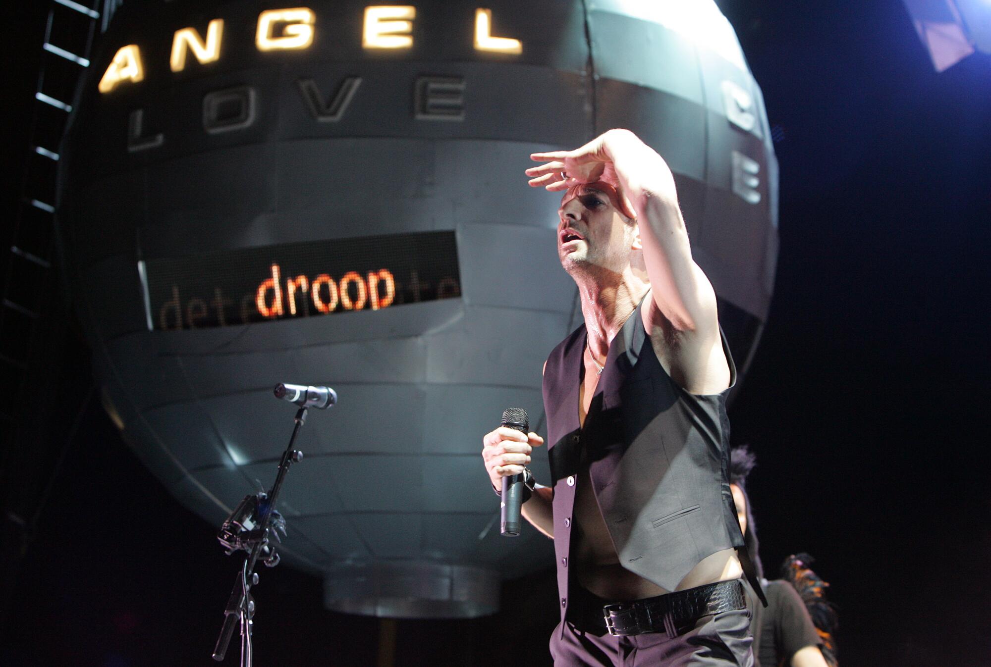 A man wears a black vest and holds his hand above his eyes in front of a ball that says Angel Love Droop