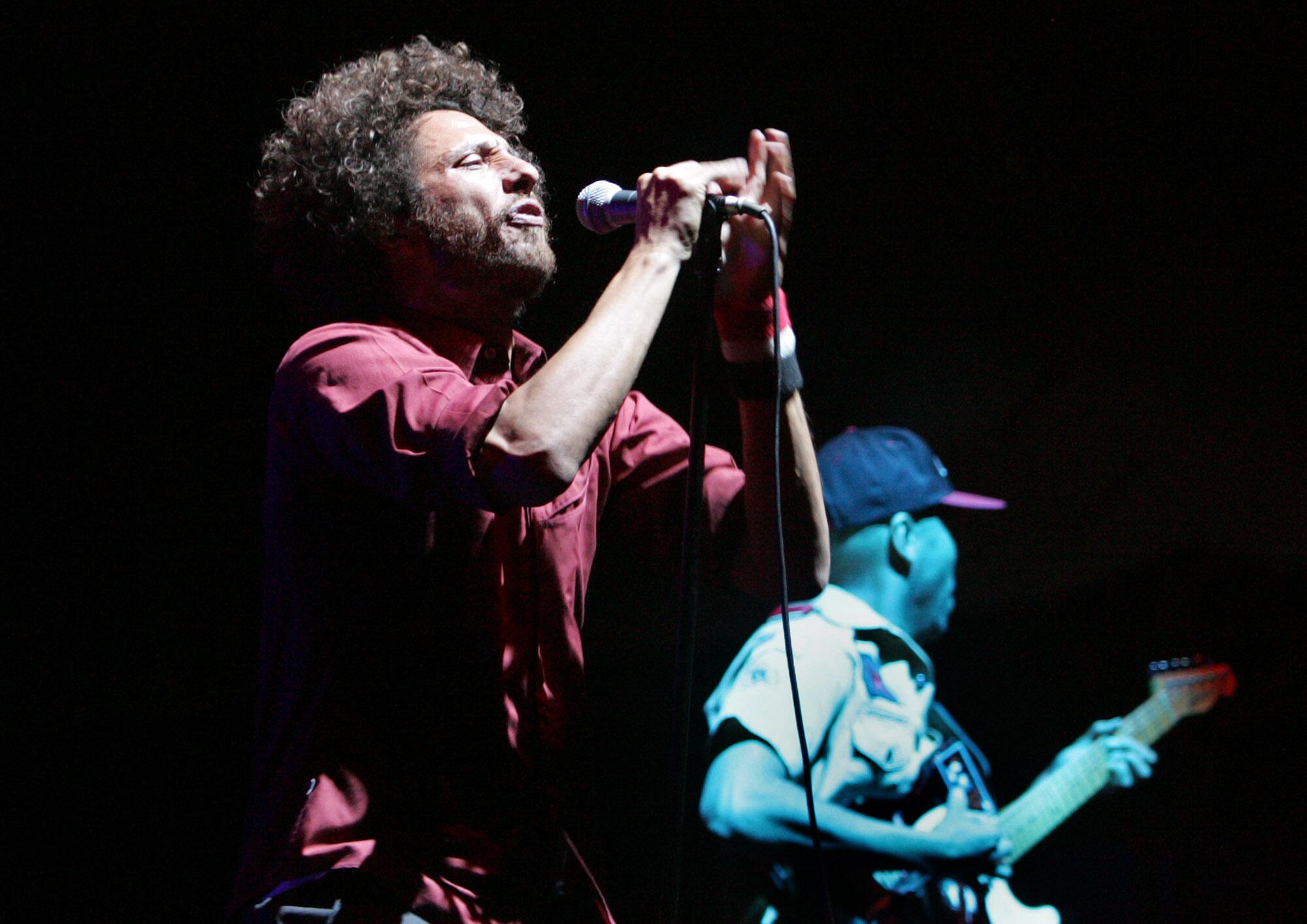 Zack de la Rocha of Rage Against The Machine sings into a microphone. Tom Morello plays guitar behind him