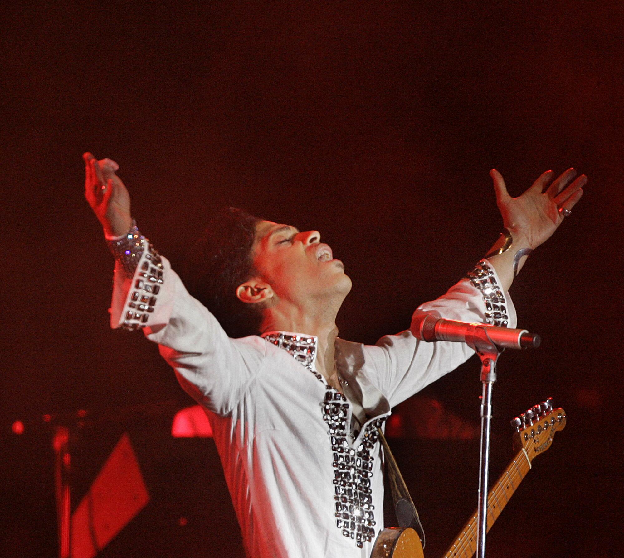 Prince stands behind a microphone with his arms outstretched and in the air