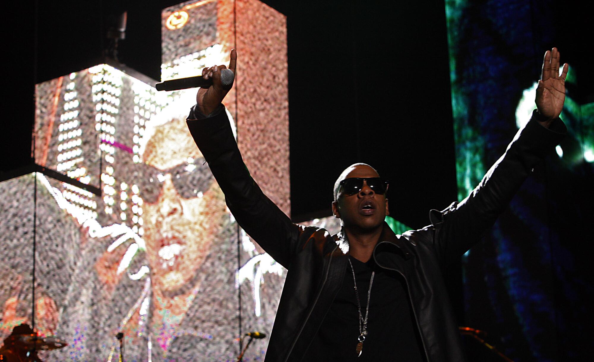  Jay Z wears sunglasses and holds a microphone in his arms stretched out above his head 
