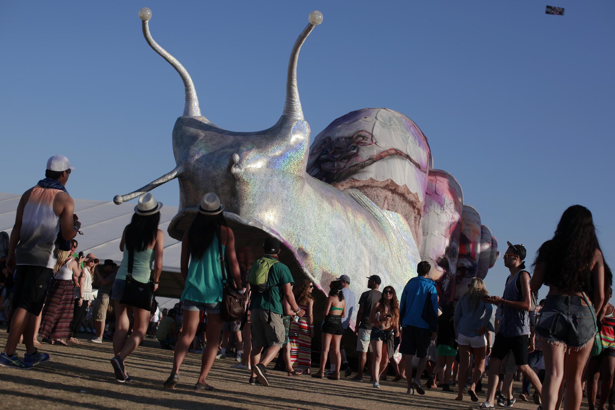 a couple of dozen festival goers walk in front of a giant snail sculpture