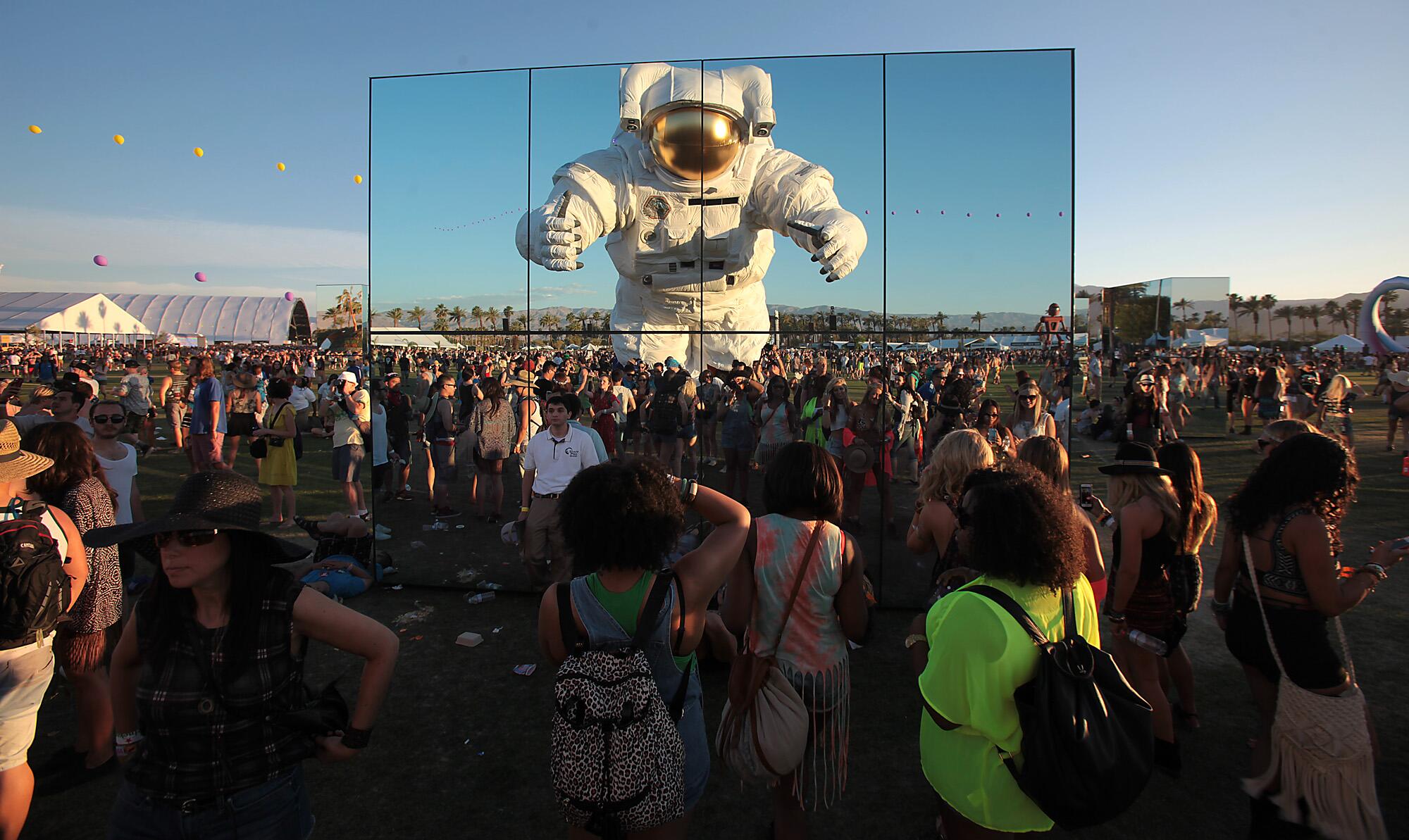A nearly 40-foot tall astronaut is reflected in a mirrored building surrounded by festival goers