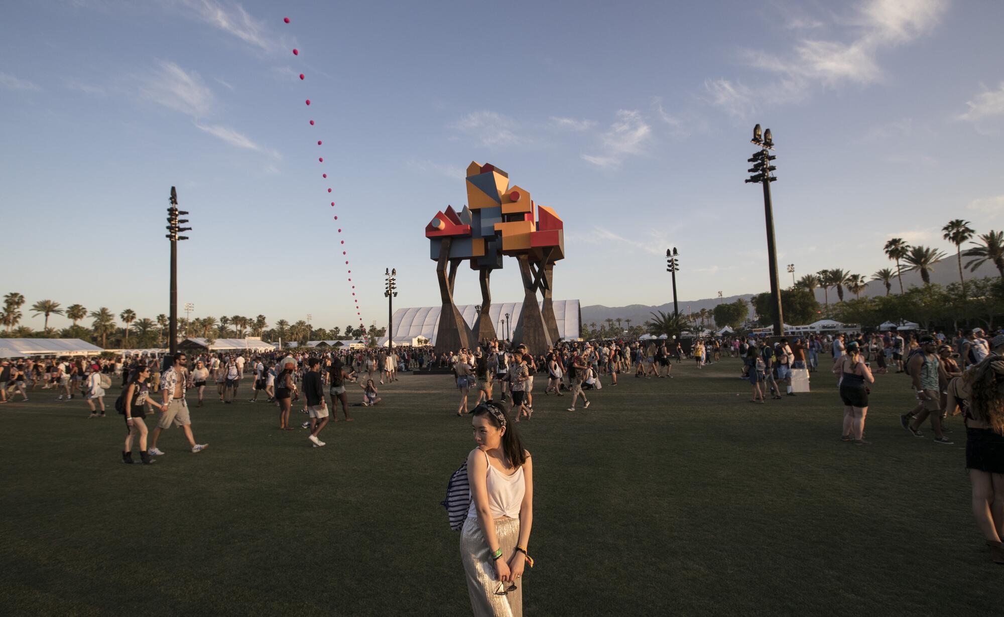 A woman poses for a photo in front of a large sculpture with people walking across a field behind her
