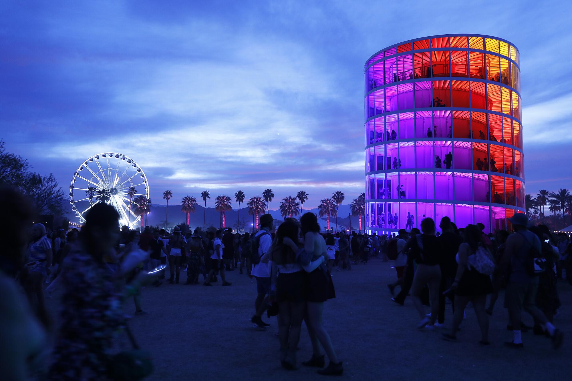 Ferris wheel and a cylinder rainbow tower with people walking by at sunset