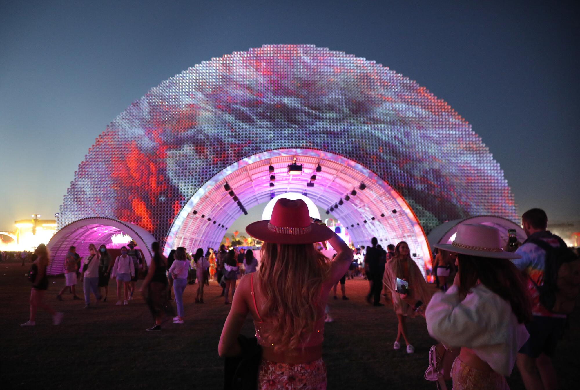 A woman with a fedora stands in front of a colorful half circle art installation at dusk