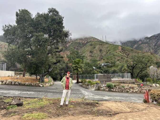 A middle aged man with light skin wearing a red baseball cap and green jacket stands at the end of a dirt lot. Behind him are mountains and a large oak tree. 