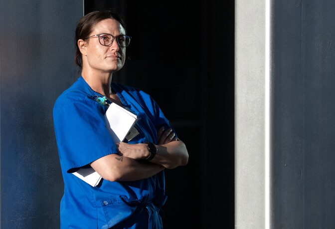 A woman with light skin tone, wearing blue scrubs and glasses, poses for a photo near a doorway and she looks out of frame.