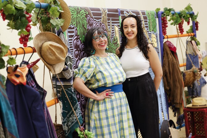 Two women stand side by side next to a collection of theater props. Both are medium light skin tone. One wears a patterned dress and blue-rimmed glasses, the other wears a white shirt and black pants.