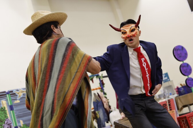 A man in a navy blue suit appears to confront a man in a striped poncho. The suited man wears a bright red tie that says "Politician" and wears a mask with horns.