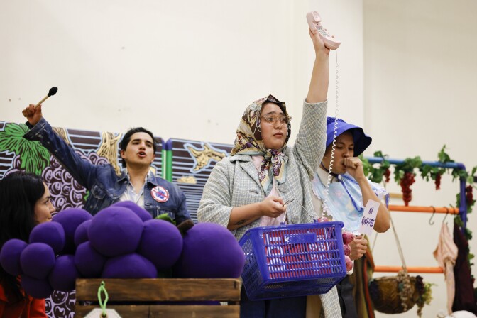 A woman with a medium-light skin tone and a scarf over her head holds a phone on the air, while holding a blue basket in her other hand.