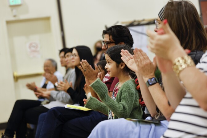 A small girl with medium-light skin tone sits in the middle of a row of people clapping their hands.