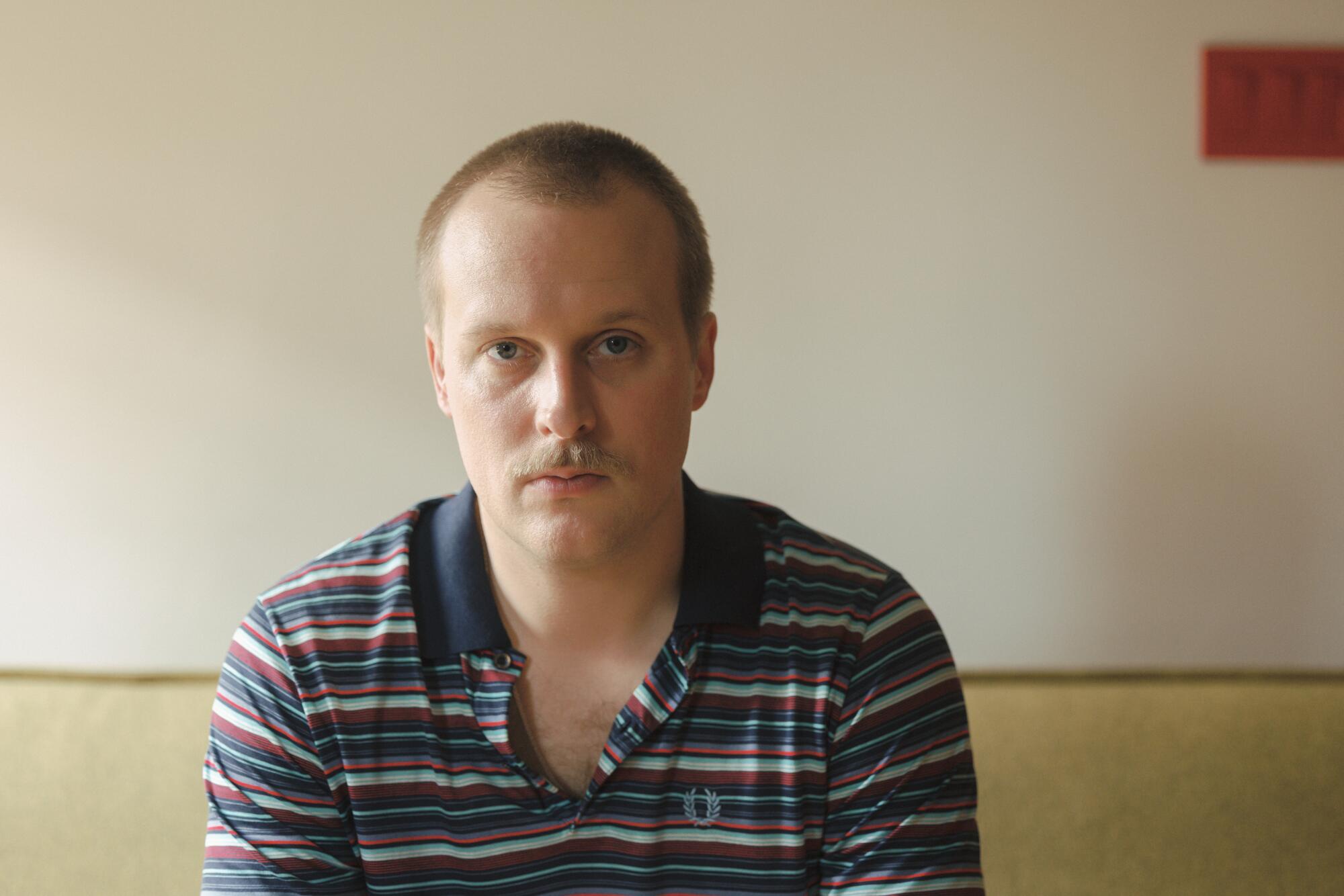 A man in a polo shirt sits on a couch and stares at the lens.
