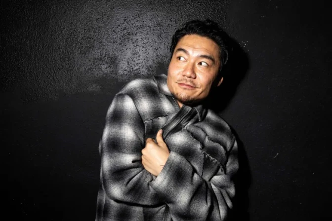 Rapper and actor Dumbfoundead (Jonathan Park), an Asian man with medium skin tone, wearing a black and white flannel jacket, poses for a photo while gripping his jacket, grinning, and looking to his right while standing in front of a black wall.
