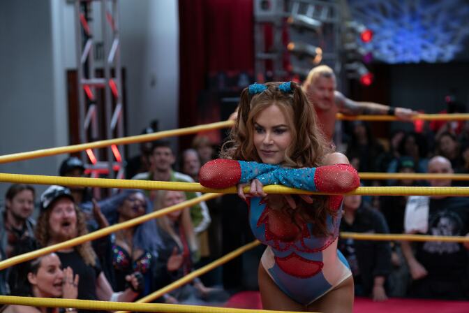 A woman stands in a wrestling ring with her arms folded on the ropes and her head on her arms. She has long red hair with pig tails on top of her head. Her costume is blue, red and white spandex and mesh. Behind her a male wrester and fans are visible but blurry.