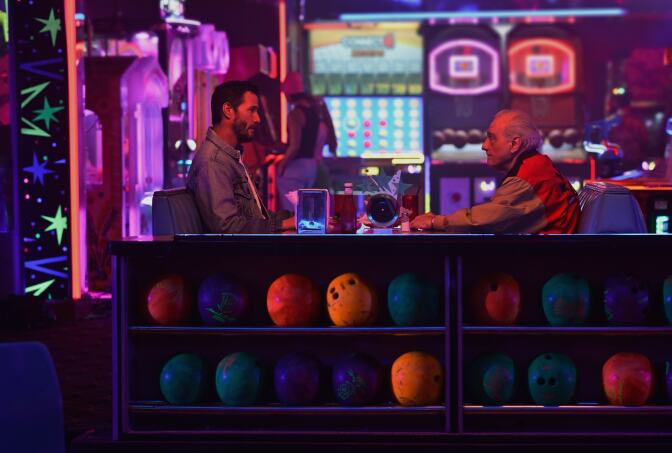 Two white men, one middle aged one older and balding, sit facing each other in a bowling alley booth. Behind them are arcade games and in front of them are rows of bowling balls. Everything is illuminated by black light so bright colors pop.