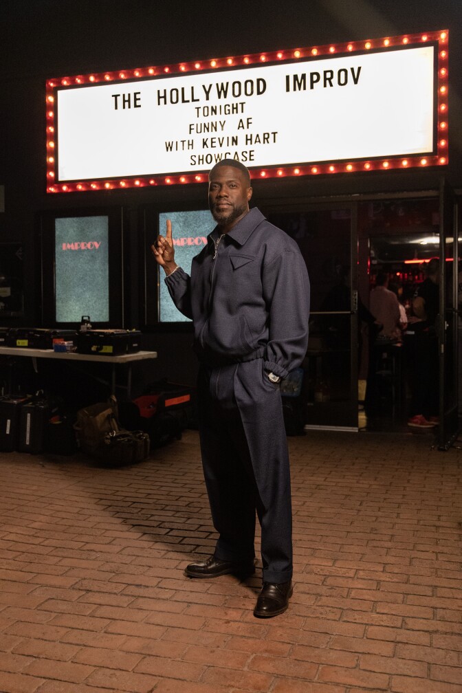 A Black man (Kevin Hart) in a black jacket and pants stands in front of a white marquee sign with red light bulbs around the edge that reads "The Hollywood Improv, Tonight, Funny AF, With Kevin Hart, Showcase." He has one hand in his pocket and one pointing up to the sign.