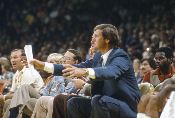 A man wearing a blue suit and carrying a rolled up piece of paper in his right hand gestures. He is surrounded by people who are looking forward.