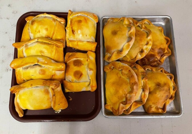A range of empanadas sit on a two metal trays; the selection on the left are rectangular in shape; the ones on the right have the more traditional half moon shape.