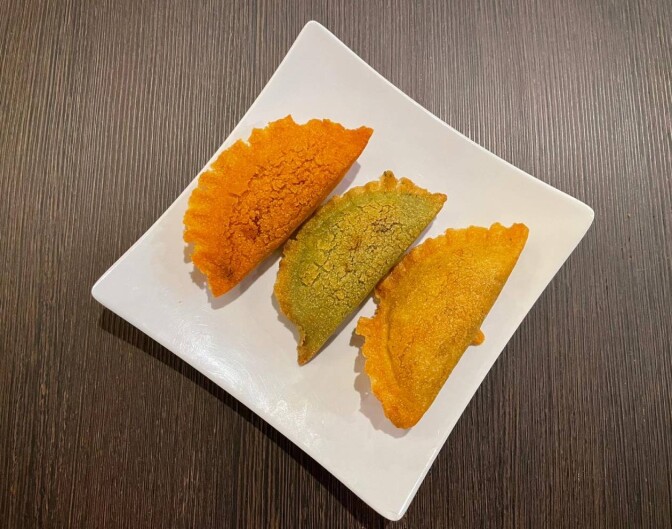A white plate sits on a wooden table. On it sit three empanadas, one slightle orange, one more yellow and one more green.
