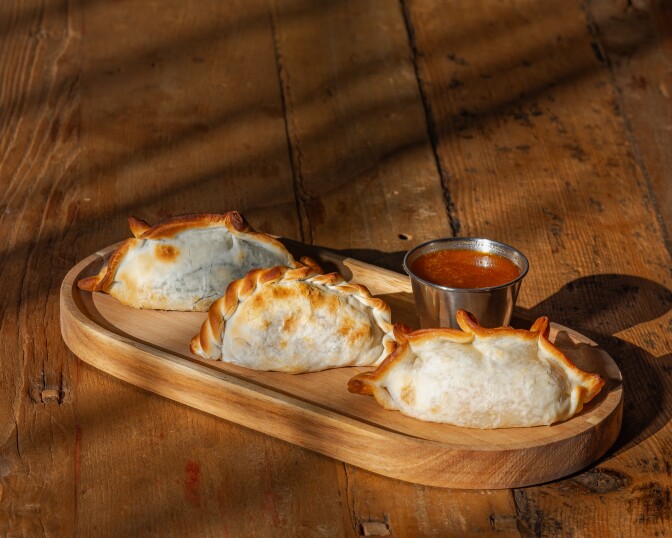 Three attractive empanadas sit on a wooden oval platter, with a metal container of tomato ketchup on the side.