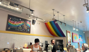 A coffee shop counter with two baristas serving drinks, a rainbow flag draped from the ceiling, colorful artwork on the walls, and a customer holding a cup.