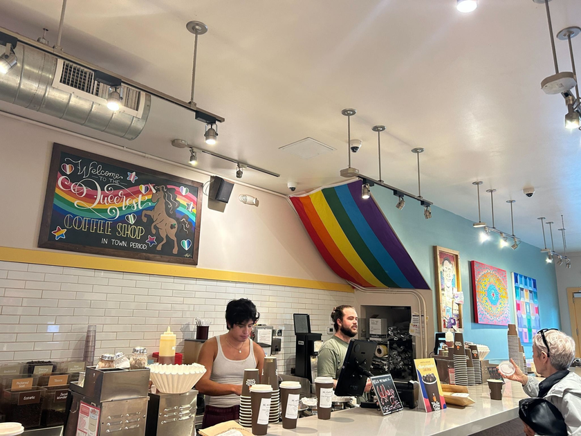 A coffee shop counter with two baristas serving drinks, a rainbow flag draped from the ceiling, colorful artwork on the walls, and a customer holding a cup.