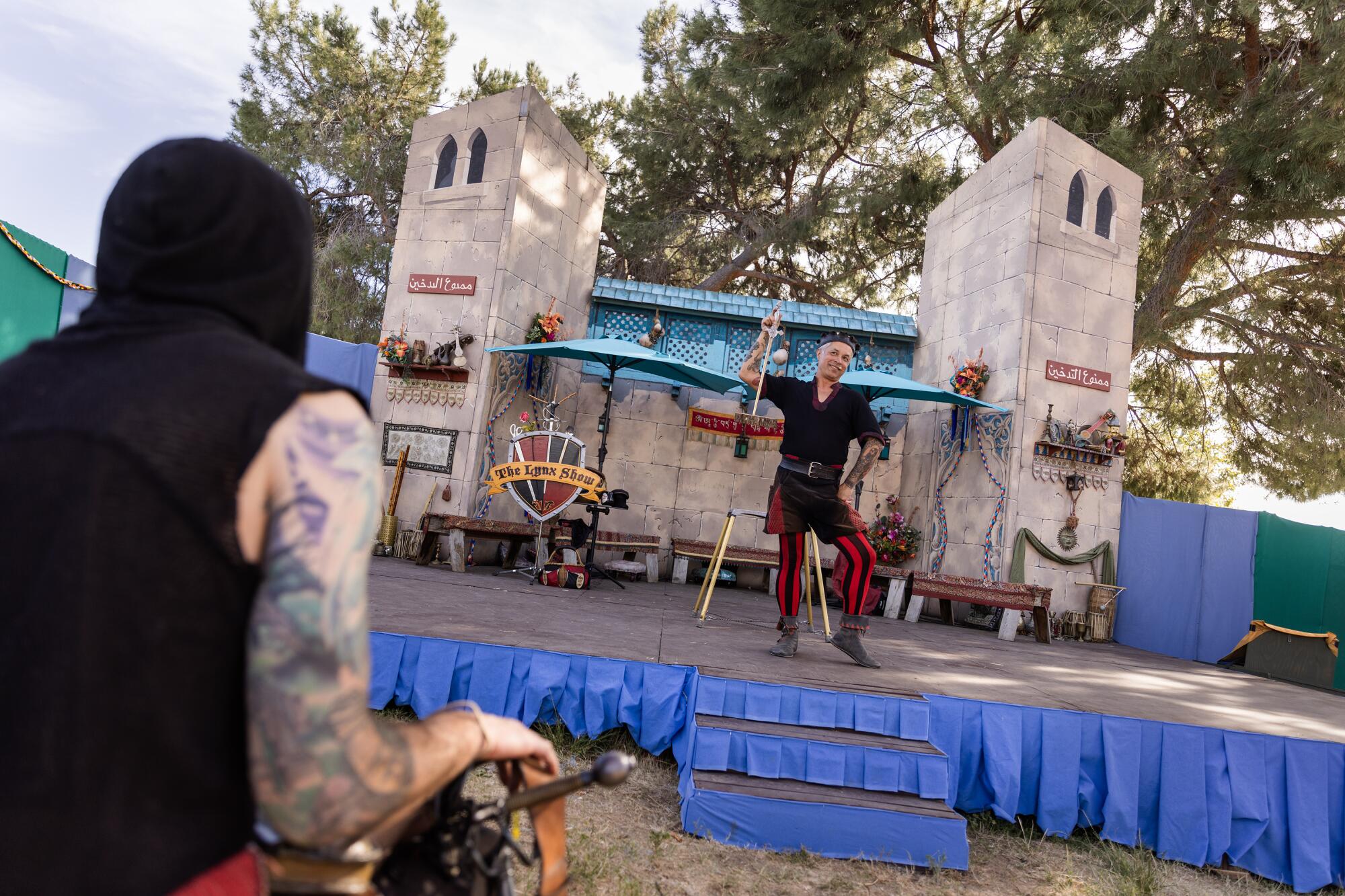 A performer in a black tunic and red and black striped pants holds a short sword while standing on a medieval themed stage