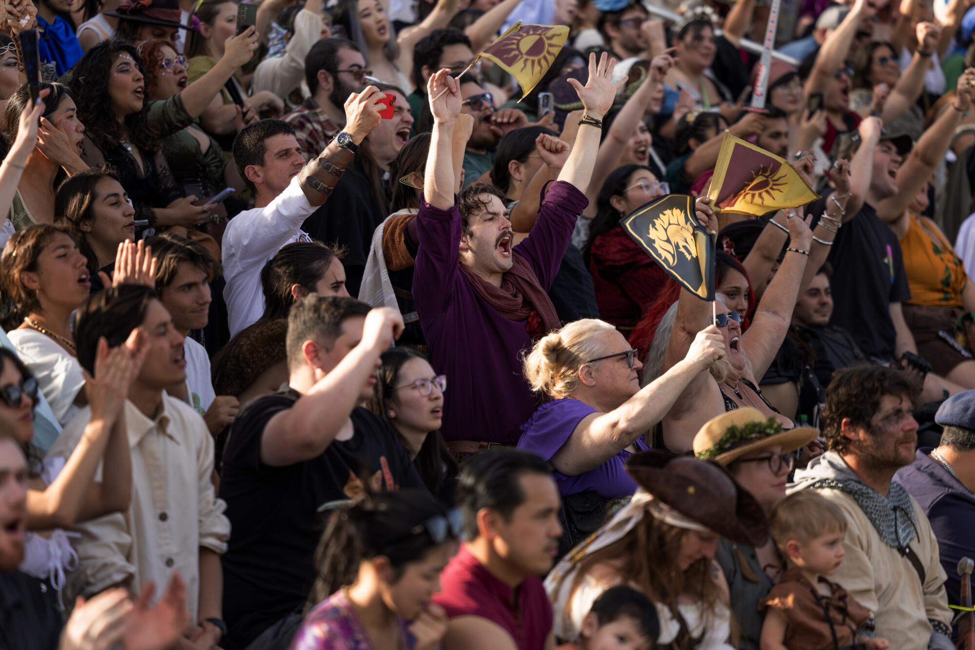 A boisterous crowd of people, some donning medieval themed costumes, others holding black and gold and crimson and gold flags