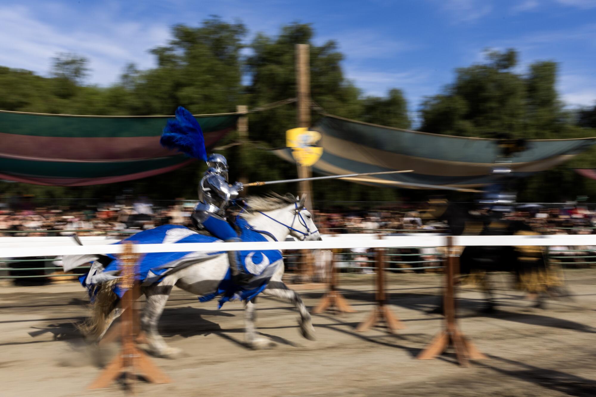 A knight with a large blue plume on his helmet rides a white horse as he charges ahead with a long thin lance